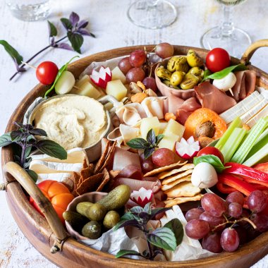 Eine reichlich gefüllte Brotzeitplatte mit Käse, Schinken, Dips, Gemüse, Obst und Cracker. Im Hintergrund stehen Wein und Wassergläser und noch mehr Cracker.