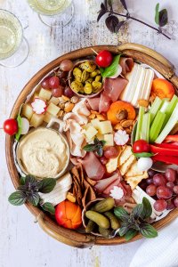 Eine reichlich gefüllte Brotzeitplatte mit Käse, Schinken, Dips, Gemüse, Obst und Cracker. Im Bildrand stehen Wein- und Wassergläser.