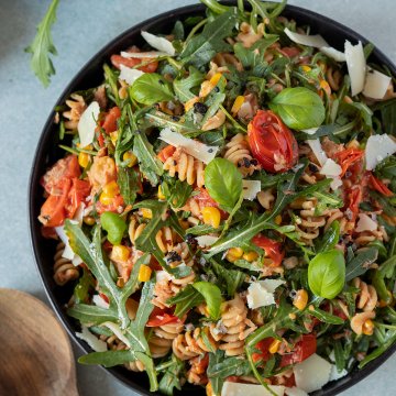 Nahaufnahme von einem Teller Nudelsalat mit Thunfisch, Tomaten, Rucola und Parmesan, Salatbesteckt und eine Schüssel mit Parmesan im Bildrand.