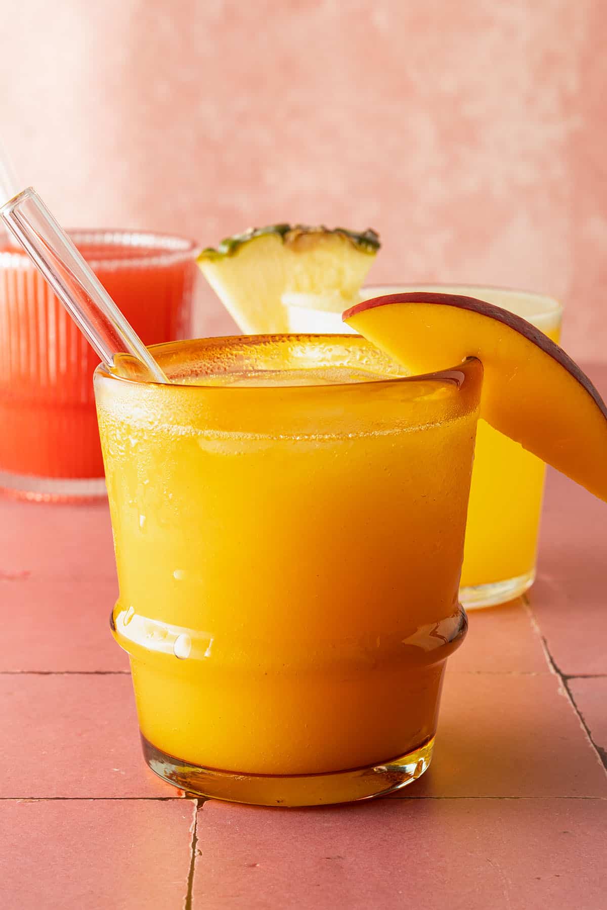 Glas mit gelber Agua Fresca mit Mangospalte und Glasstrohhalm, im Hintergrund weitere Agua Fresca Sorten.