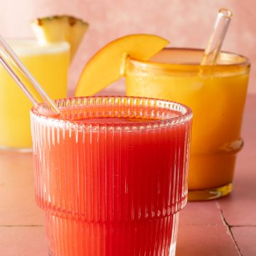 Drei Gläser Agua Fresca in verschiedenen Farben, vorne rote Erdbeer Agua Fresca mit Glasstrohhalm.