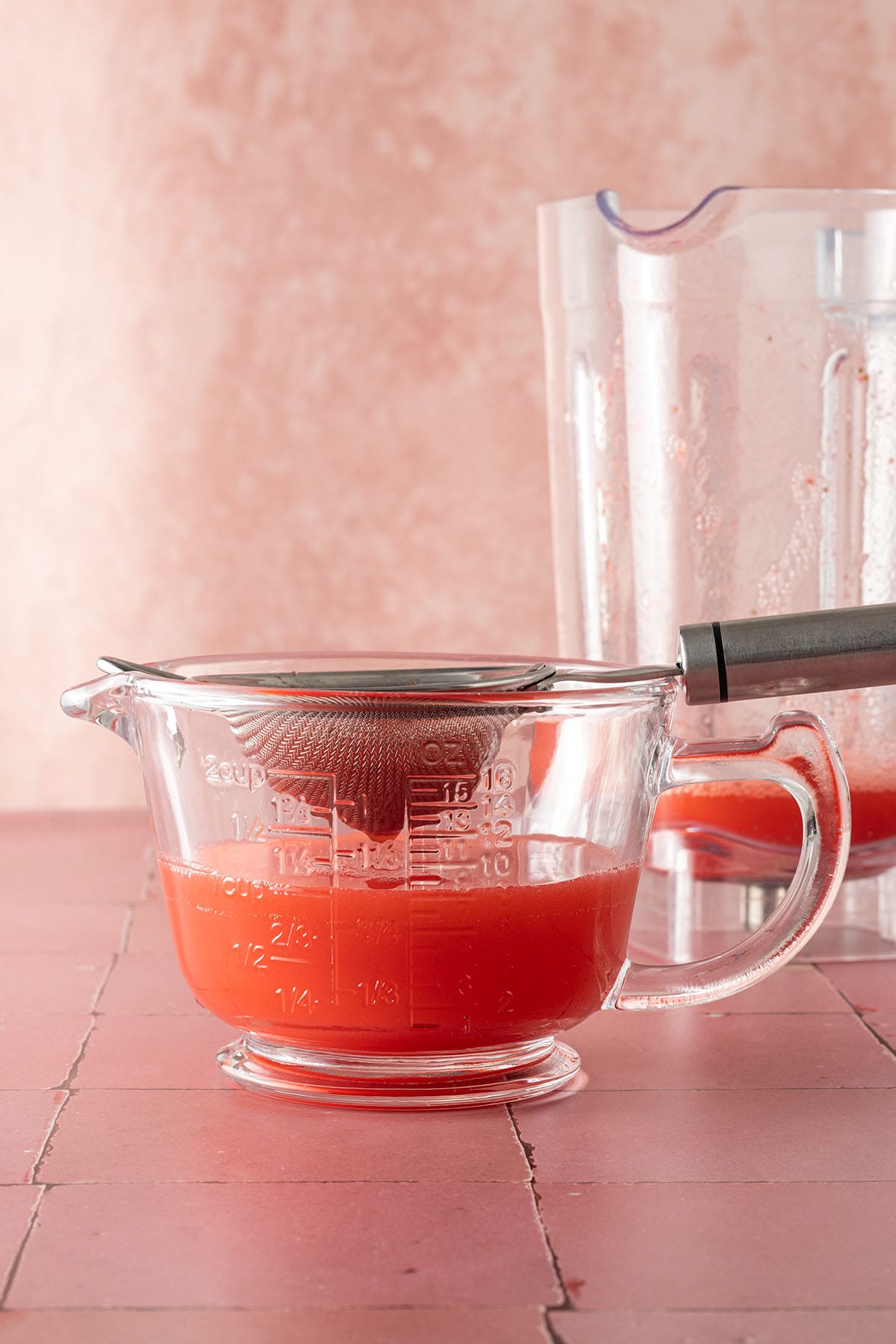 Erdbeer-Agua-Fresca wird durch ein feines Sieb in einen Messbecher gegossen, im Hintergrund der Mixerbehälter.