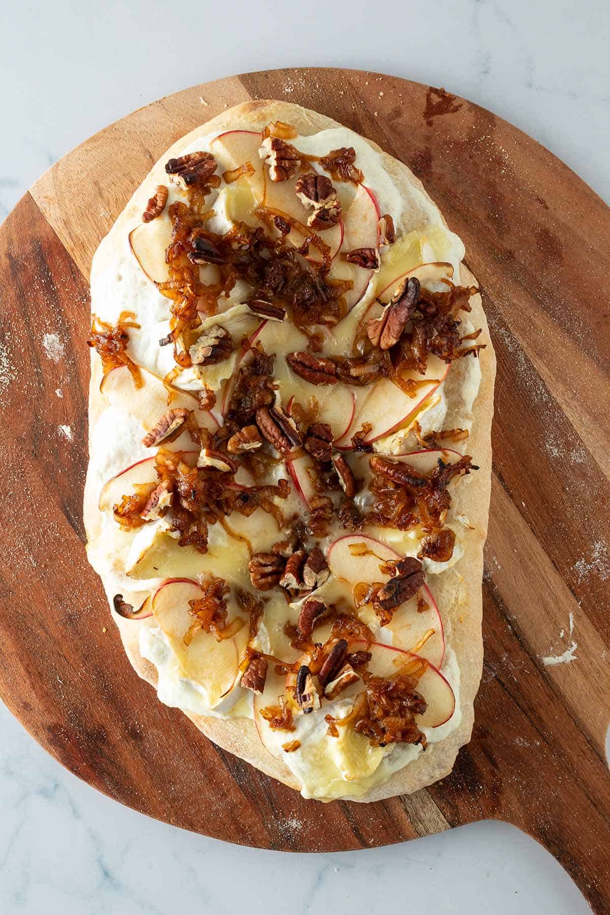 Pinsa Bianca nach dem Backen mit Ricotta, Brie, Apfelscheiben, gebratenen Schalotten und Pekann&uuml;ssen auf einem Holzbrett.