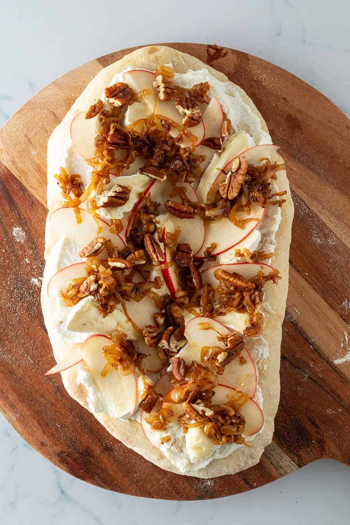 Pinsa Bianca vor dem Backen mit Ricotta, Brie, d&uuml;nnen Apfelscheiben, Schalotten und Pekann&uuml;ssen belegt.