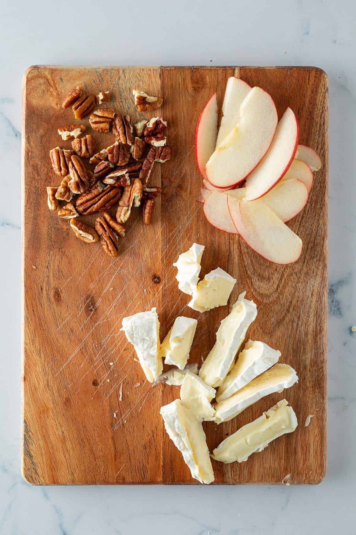 Vorbereitete Zutaten f&uuml;r Pinsa Bianca: in Scheiben geschnittener Camembert, Apfel und grob zerkleinerte N&uuml;sse auf einem Brett.