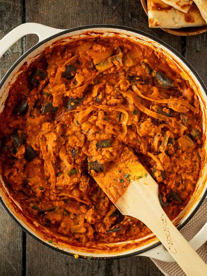 Auberginen Curry mit Linsen und Zwiebeln in einer cremigen Tomatensauce in einer großen Pfanne auf einem rustikalen Holzuntergrund, im Bildrand Naan.