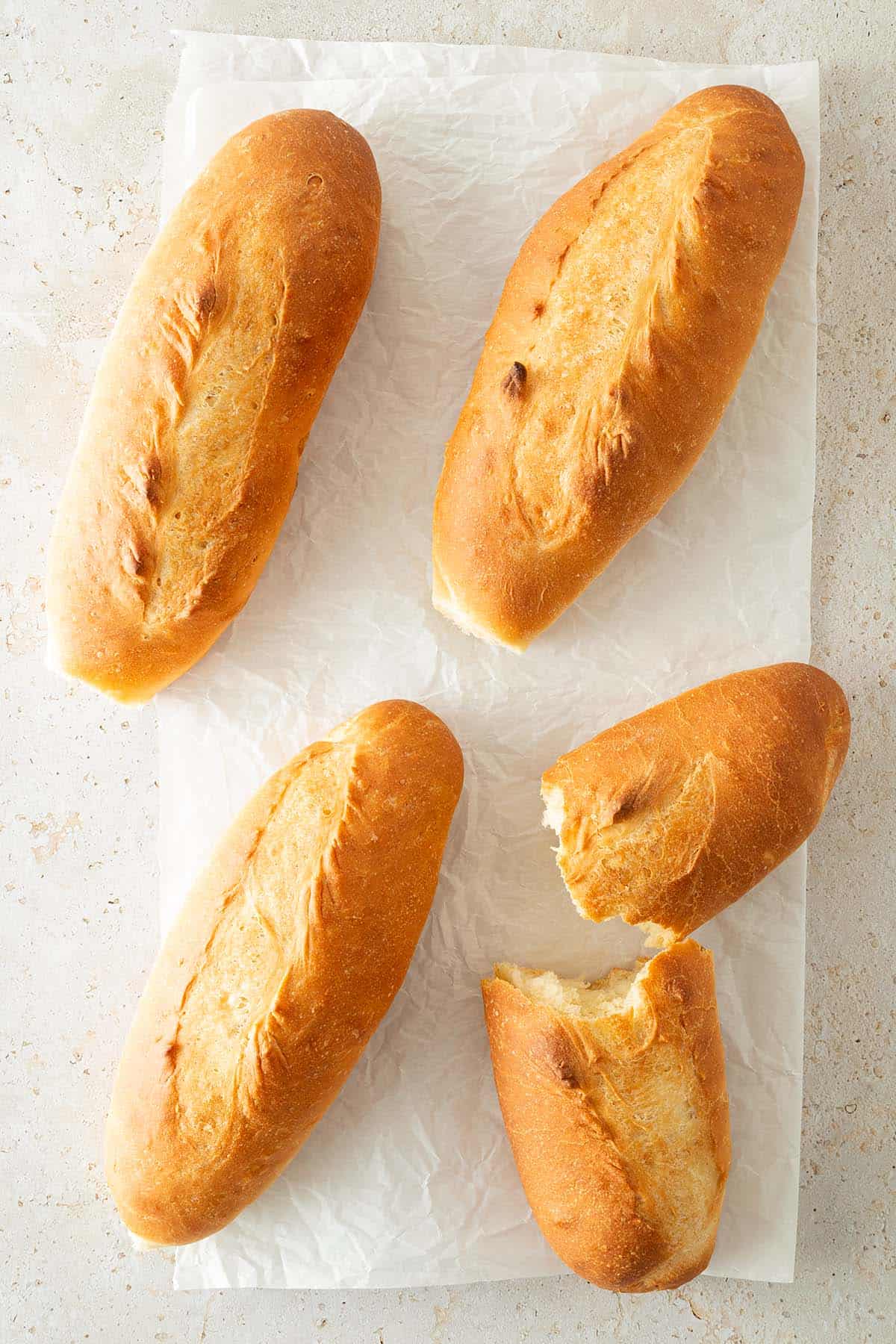 Vier goldbraune, frisch gebackene Banh-Mi-Baguettes auf Backpapier, eines aufgebrochen.