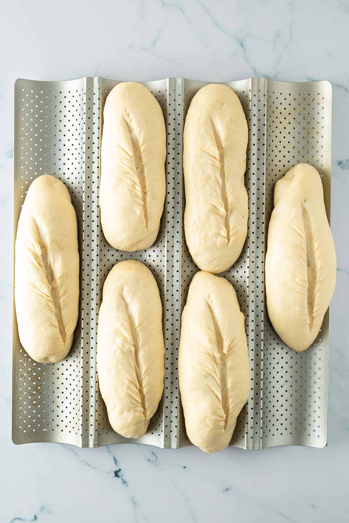 Sechs geformte Baguette-Teiglinge mit Einschnitten auf perforiertem Baguette-Blech vor dem Backen.