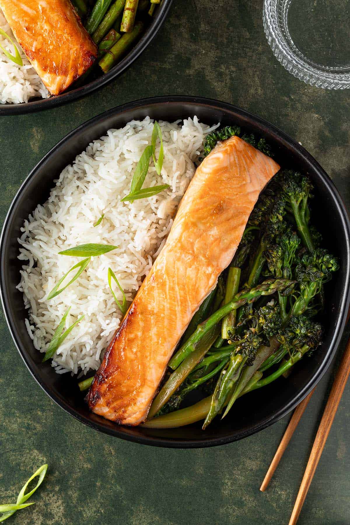 Bowl mit gebratenem Lachs, Reis und asiatischem Gem&uuml;se auf dunkelgr&uuml;nem Untergrund, Deko und zweite Sch&uuml;ssel im Bildrand.