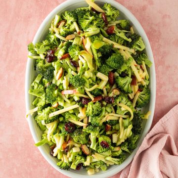 Brokkoli Salat mit Apfel, Cranberries, K&auml;se und Mandeln auf einem ovalen Teller serviert auf einem rosa Untergrund.