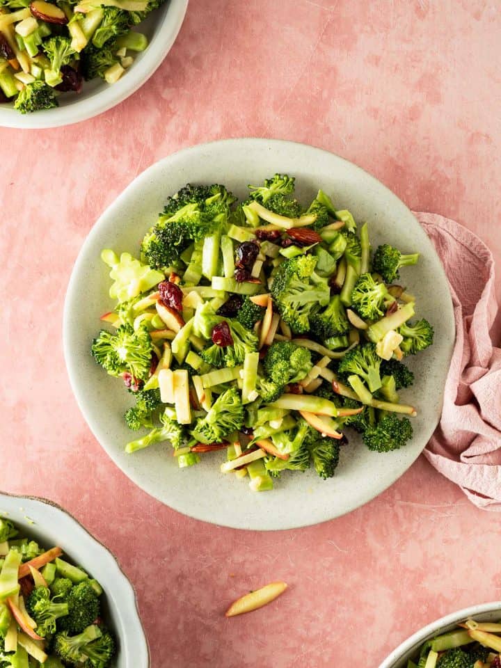 Brokkoli Salat mit Apfel, K&auml;se und Cranberries verteilt auf vier Teller, serviert auf einem rosa Untergrund.