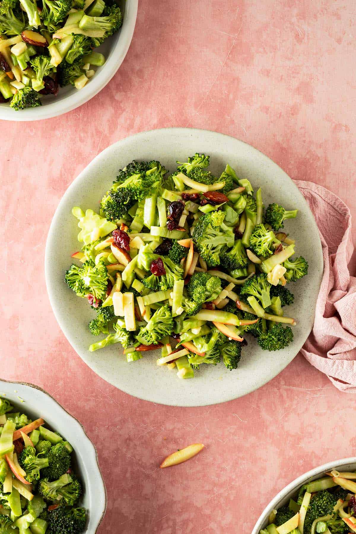 Brokkoli Salat mit Apfel, K&auml;se und Cranberries verteilt auf vier Teller, serviert auf einem rosa Untergrund.