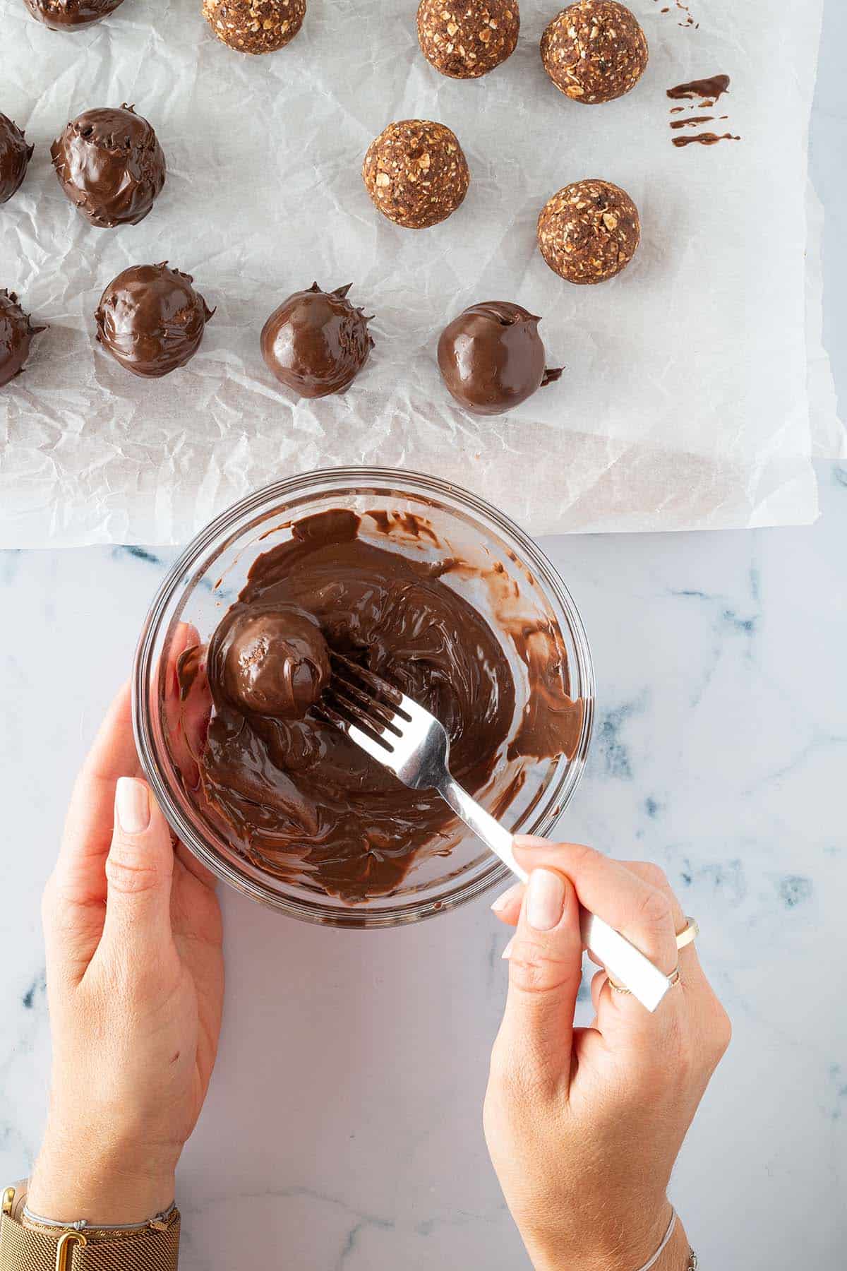 Die zu Kugel geformten Dattel-Schoko-Bällchen werden in einer Schüssel geschmolzener Schokolade gewälzt. Daneben ein Backpapier mit Schokolade überzogener Brownie Balls.