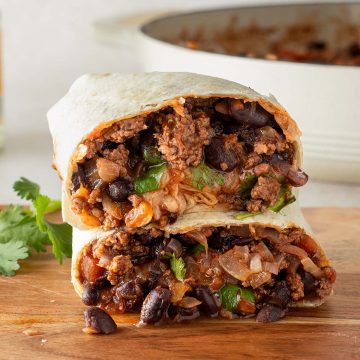Ein in der Mitte halbierter Burrito gefüllt mit einer Hackfleisch und Bohnen Pfanne auf einem Holzbrett, im Hintergrund eine Pfanne mit Chili con Carne Füllung und frische Kräuter.