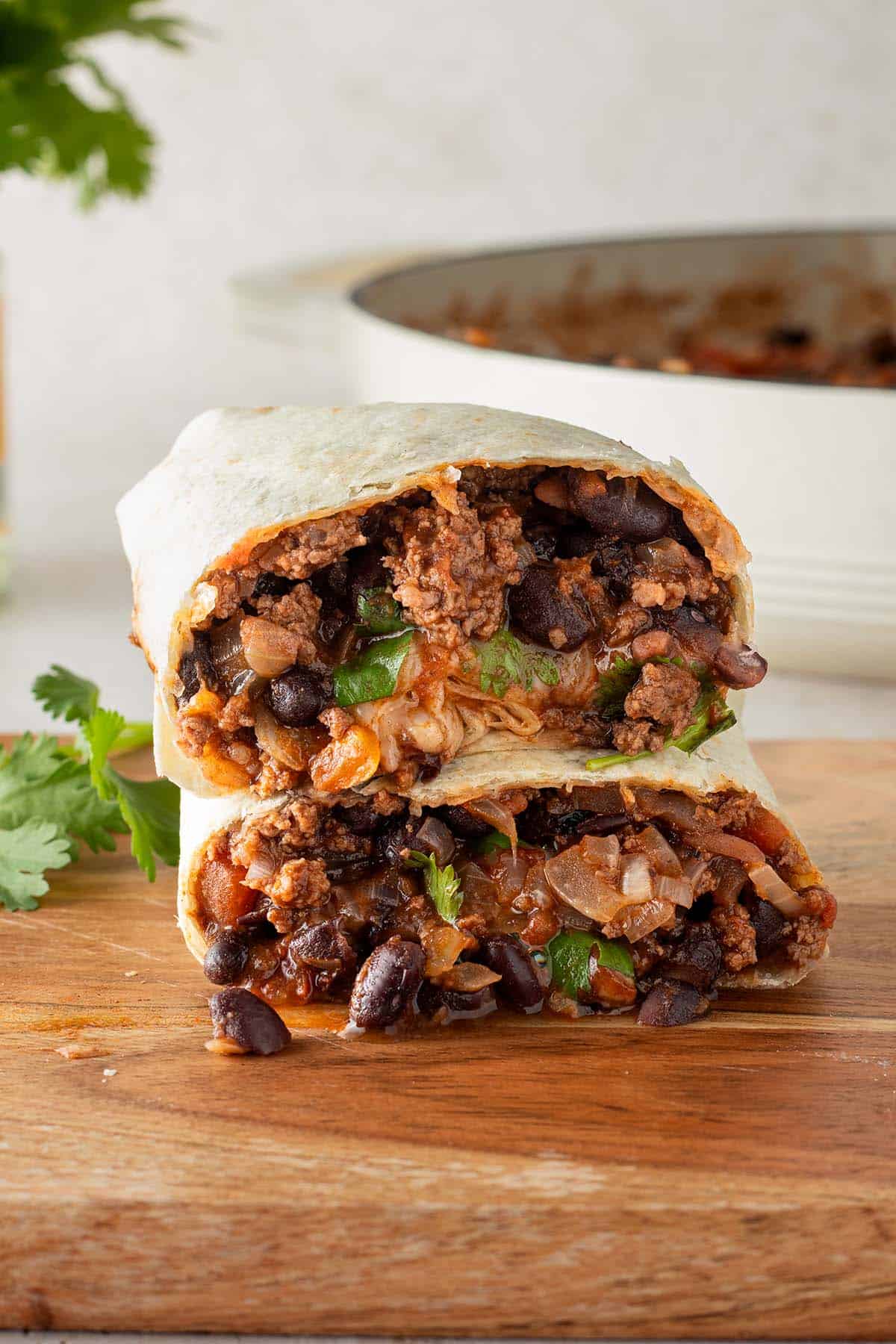 Ein in der Mitte halbierter Burrito gefüllt mit einer Hackfleisch und Bohnen Pfanne auf einem Holzbrett, im Hintergrund eine Pfanne mit Chili con Carne Füllung und frische Kräuter.
