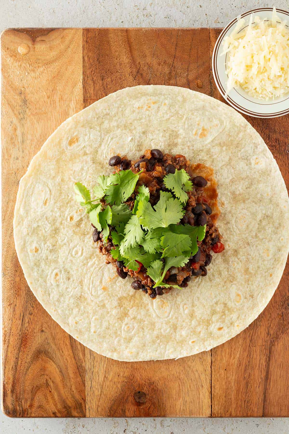 Ein Tortilla auf einem Brett mit der Hackfleisch Füllung und frischem Koriander in der Mitte. Daneben geriebener Käse auf einem kleinen Teller.
