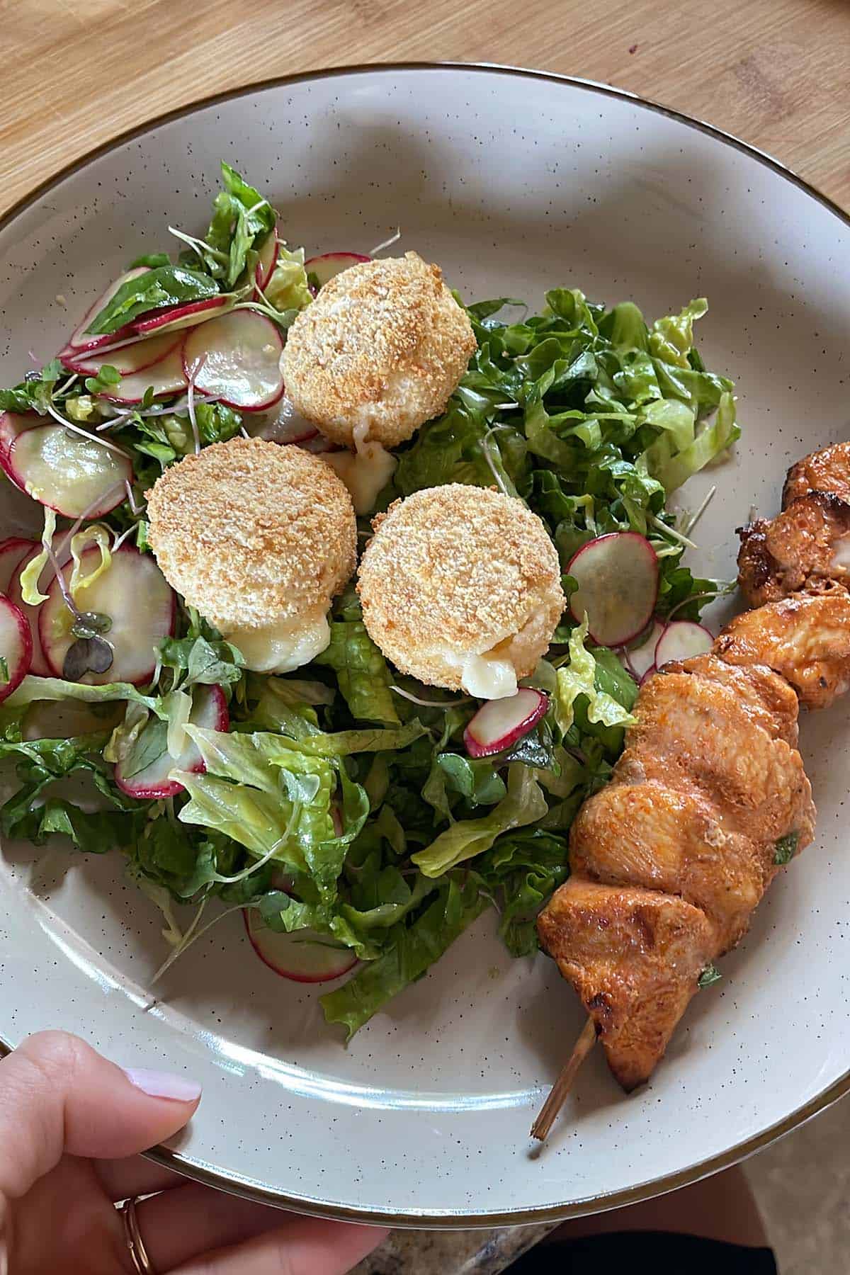 Drei kleine, im Airfryer gebackene Camemberts auf gemischten Salat auf einem Teller mit einem Hähnchenspieß.