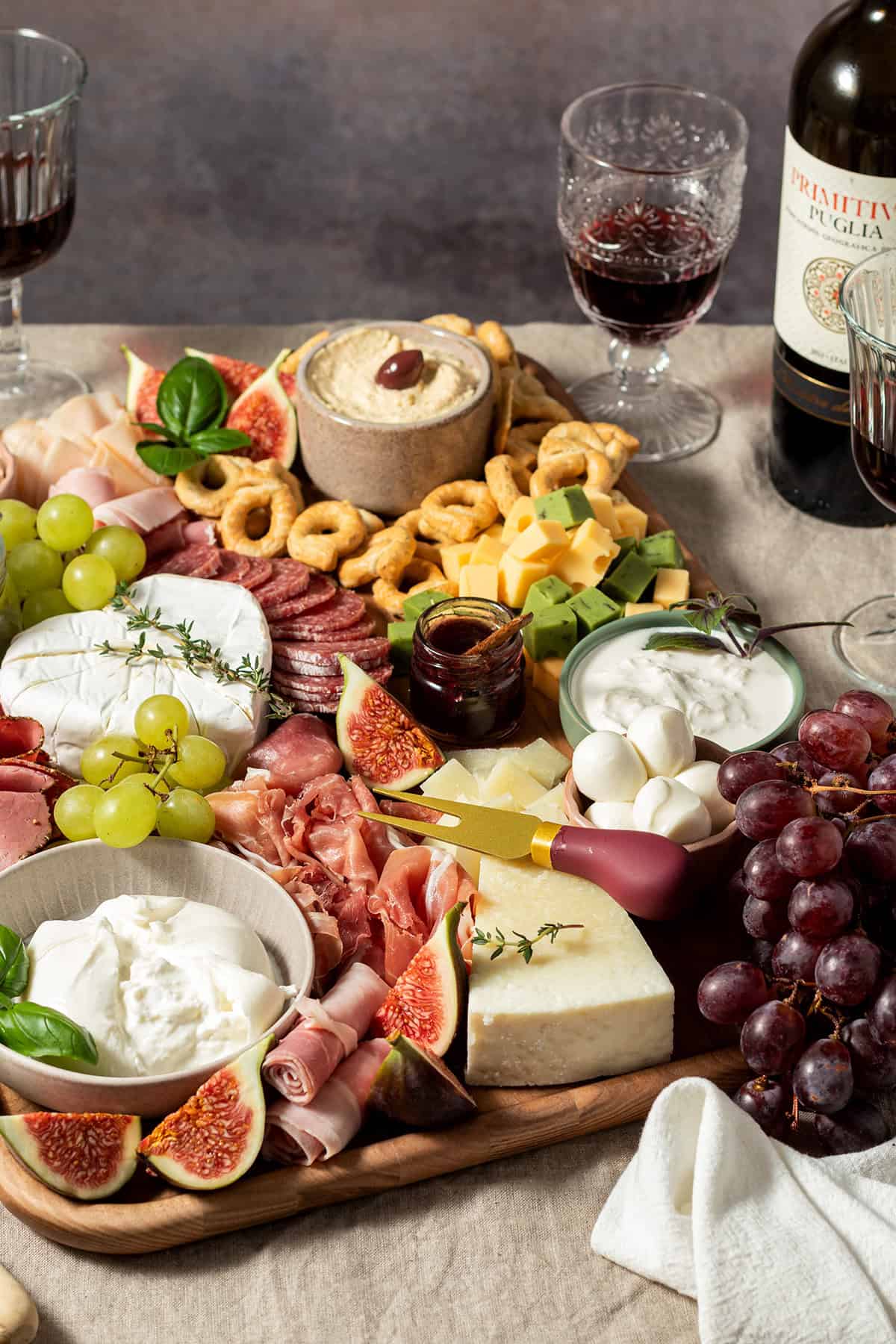 Große Charcuterie-Platte mit Käse, Wurst, Trauben, Feigen, Dips und Crackern, angerichtet auf einem Holzbrett, daneben Rotwein.