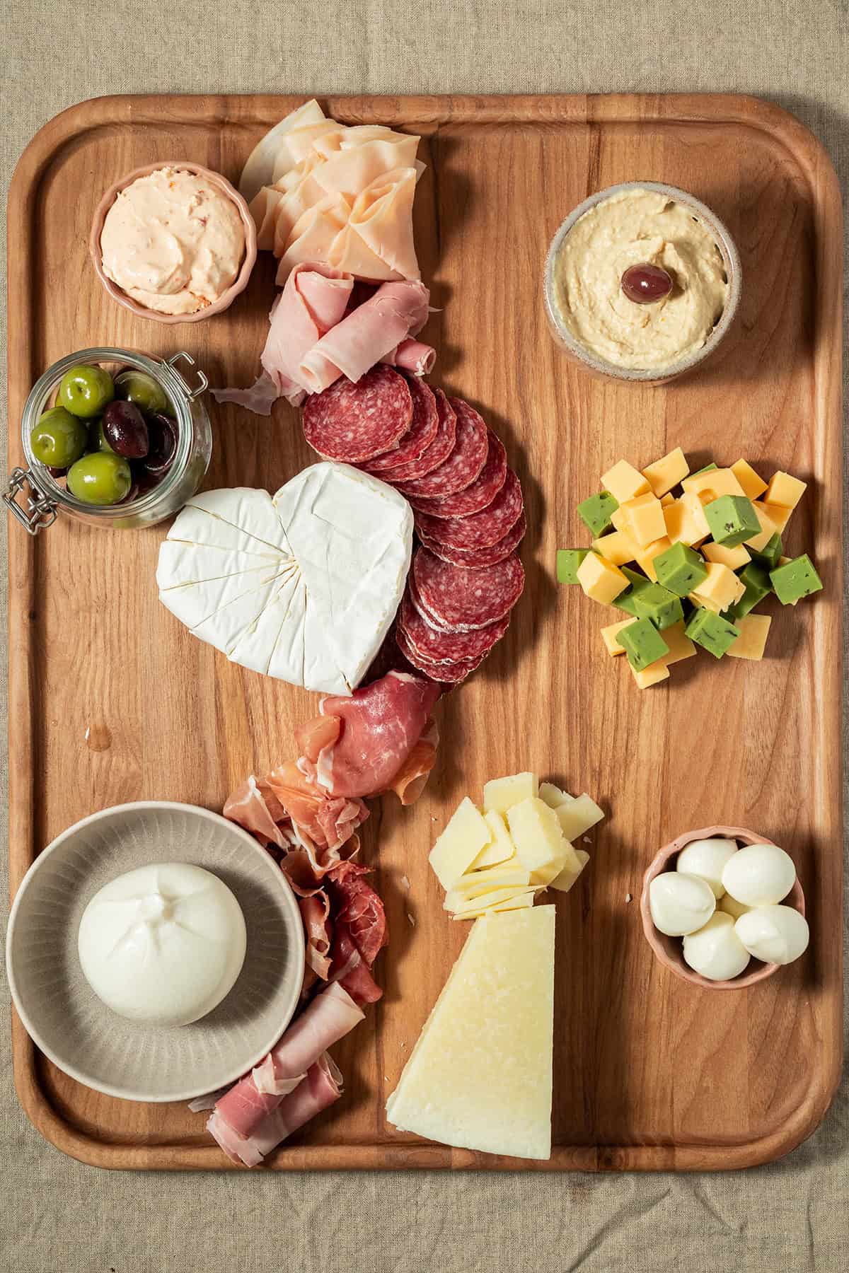 Charcuterie-Platte in der Aufbauphase mit Käse, Wurst, Trauben, Feigen, Dips und Oliven auf einem Holzbrett.