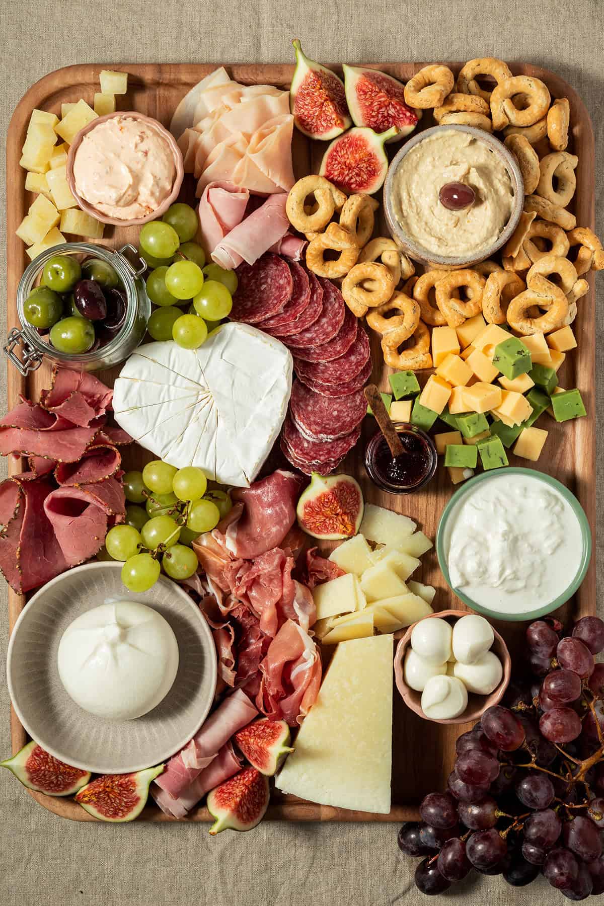 Vollständig angerichtete Charcuterie-Platte mit Käse, Wurst, Feigen, Trauben, Crackern, Dips und Antipasti auf einem Holzbrett.