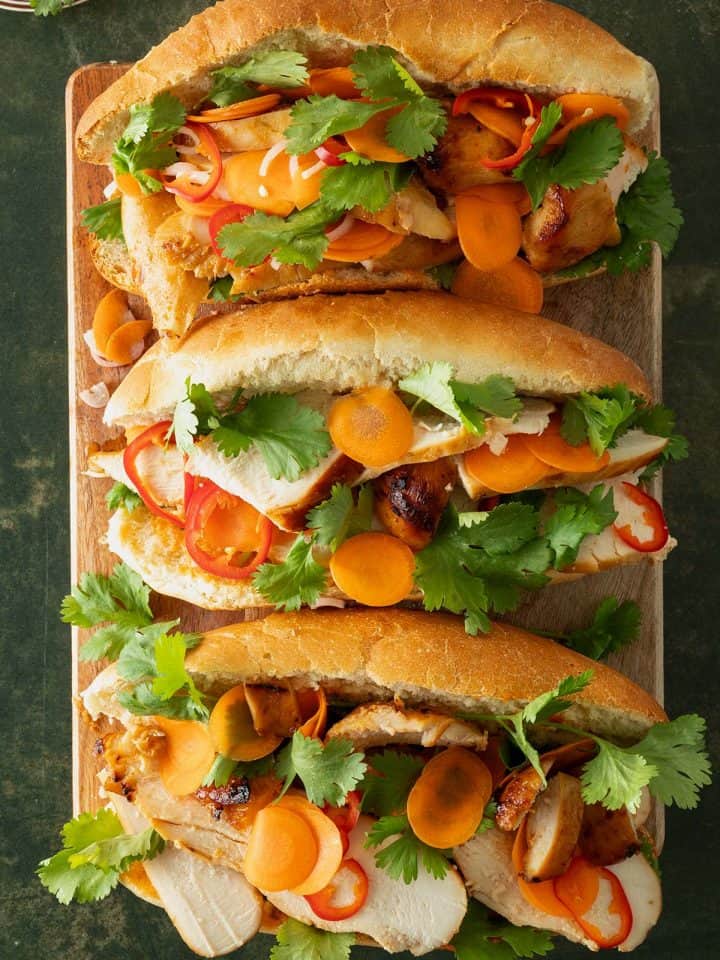 Drei Chicken Banh Mi Sandwiches mit eingelegten Karotten, Chili und frischem Koriander auf Holzbrett.