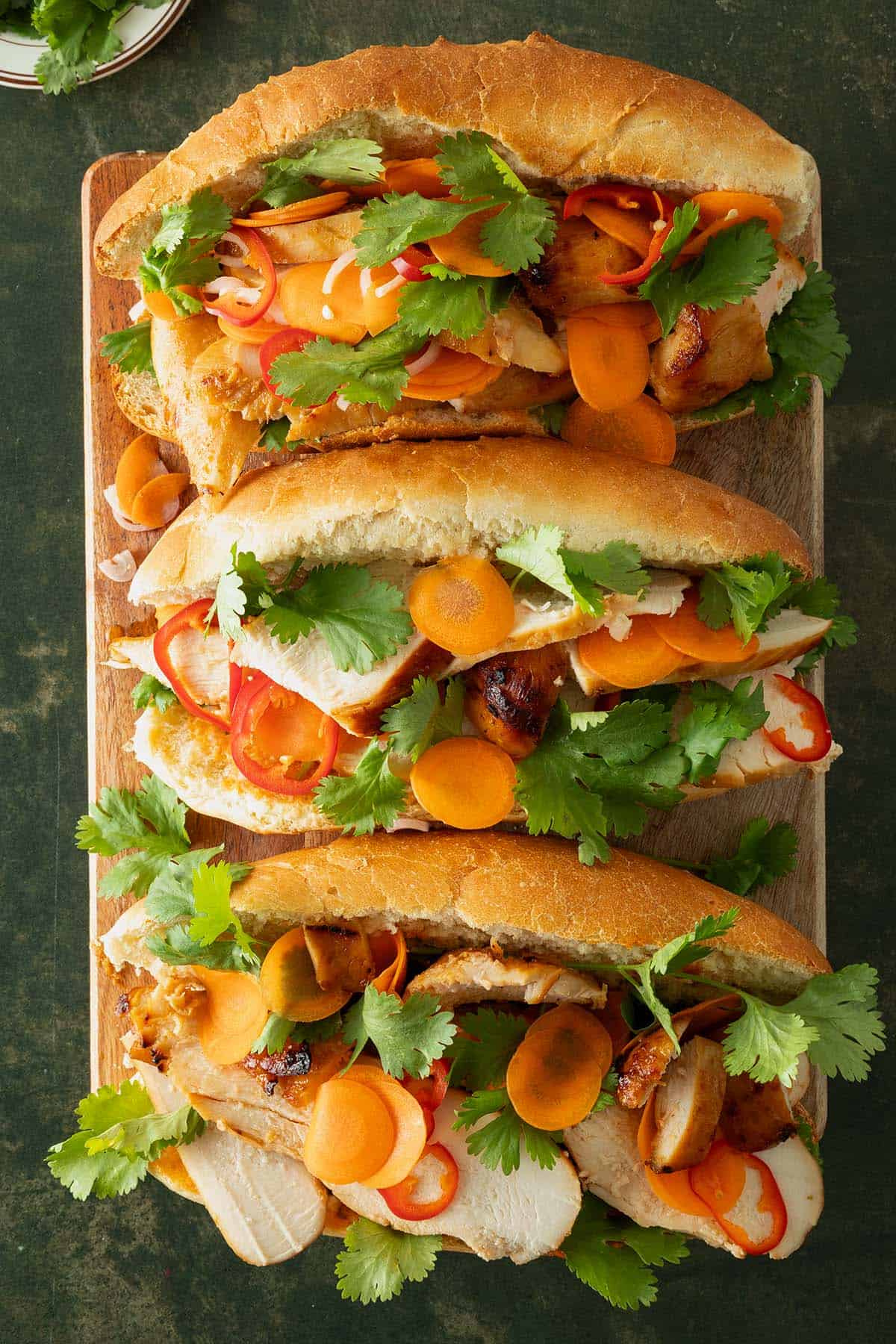 Drei Chicken Banh Mi Sandwiches mit eingelegten Karotten, Chili und frischem Koriander auf Holzbrett.
