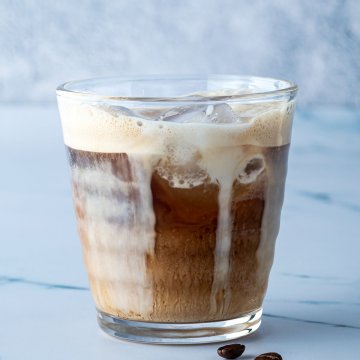 Einfache Eiskaffee aus kalt geschütteltem Espresso und einem Schuss Milch in einem Glas vor grau-blauem Hintergrund.