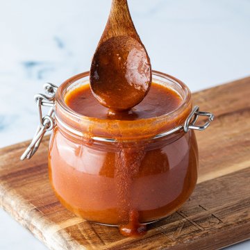 Löffel wird in ein Glas mit einer dickflüssigen Enchilada-Sauce getaucht, das auf einem Holzbrett steht.