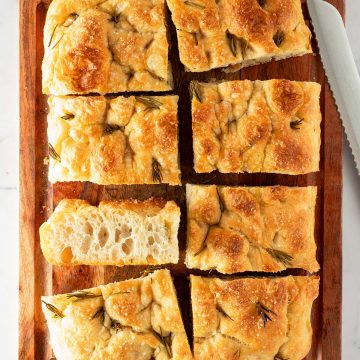 In rechteckige Stücke geschnittene Focaccia mit Rosmarin und grobem Salz auf einem Holzbrett, daneben ein Brotmesser.