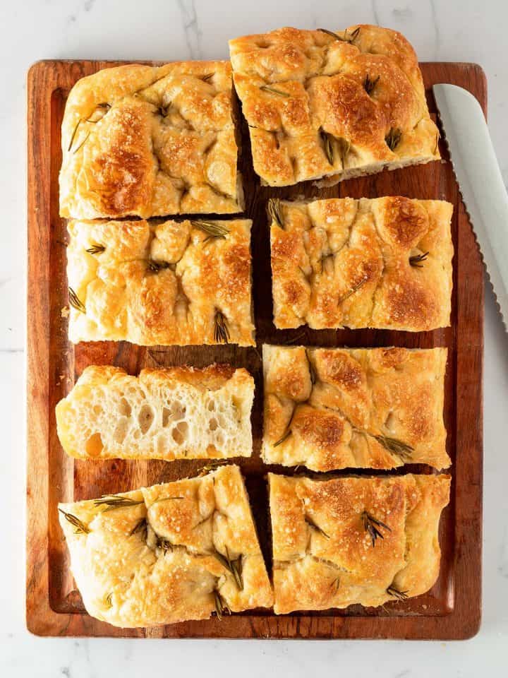 In rechteckige St&uuml;cke geschnittene Focaccia mit Rosmarin und grobem Salz auf einem Holzbrett, daneben ein Brotmesser.