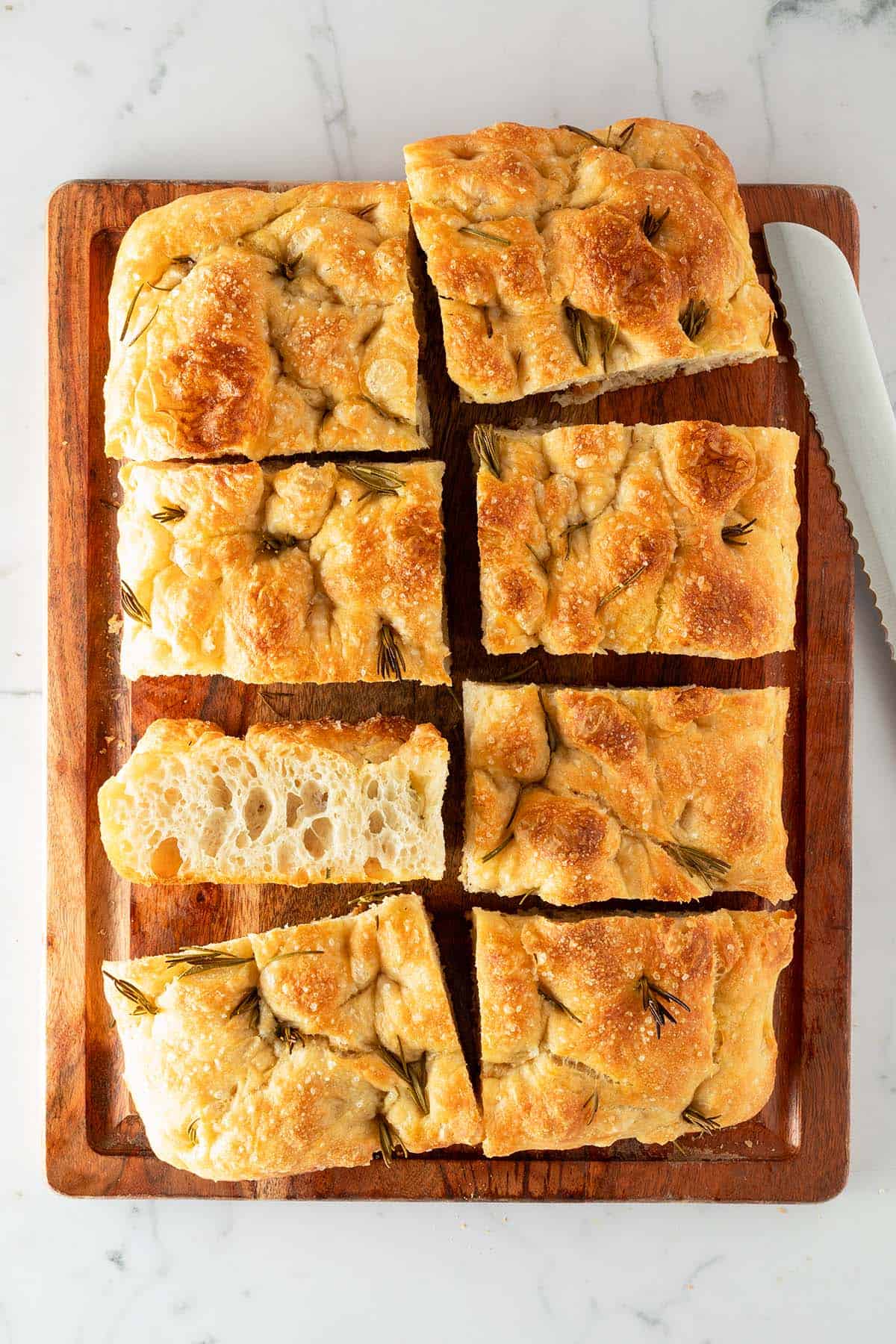 In rechteckige Stücke geschnittene Focaccia mit Rosmarin und grobem Salz auf einem Holzbrett, daneben ein Brotmesser.