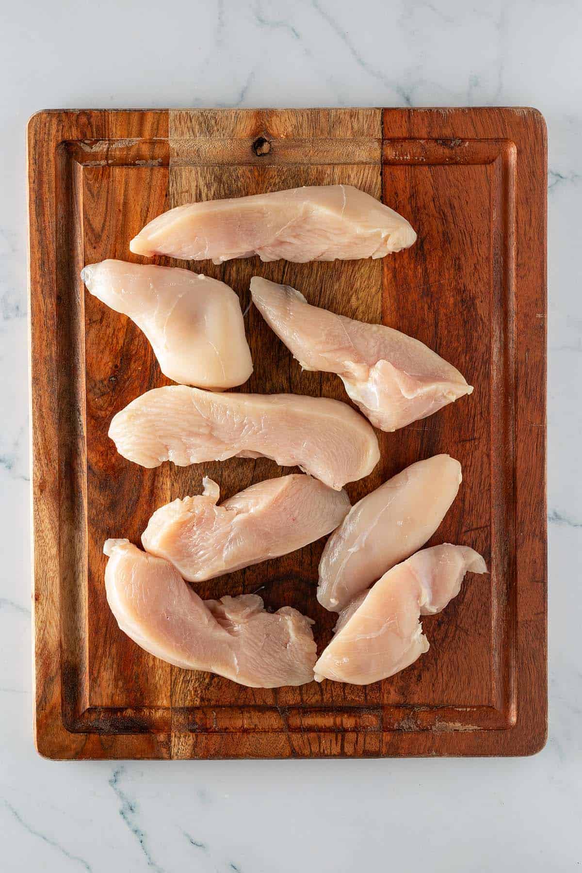 Rohe in circa 4 Zentimeter dicke Streifen geschnittene Hähnchenbrustfilets auf einem Holzbrett.