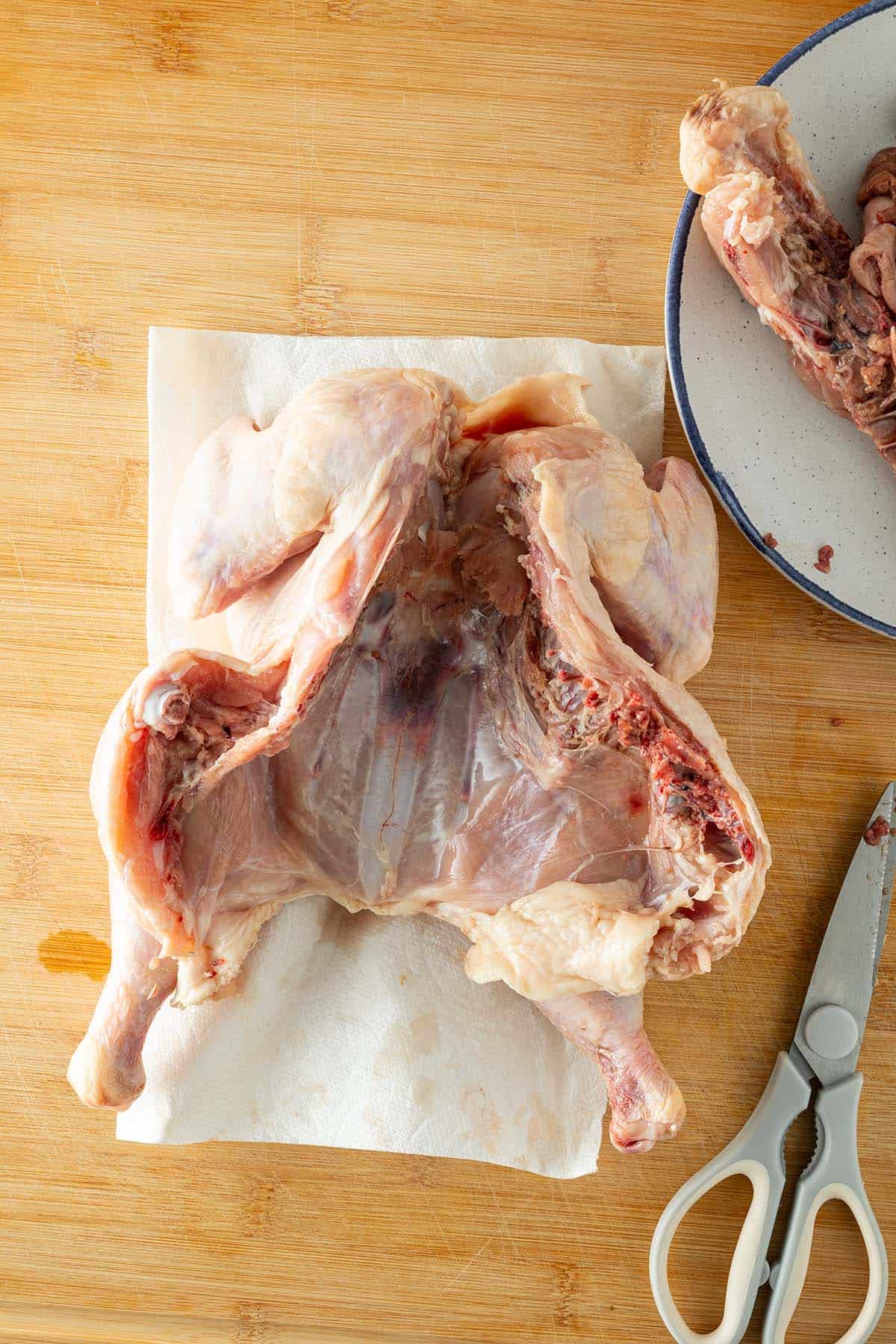 Roher Hähnchenkörper geöffnet auf einem Brett, Rückenknochen und Schere liegen daneben.