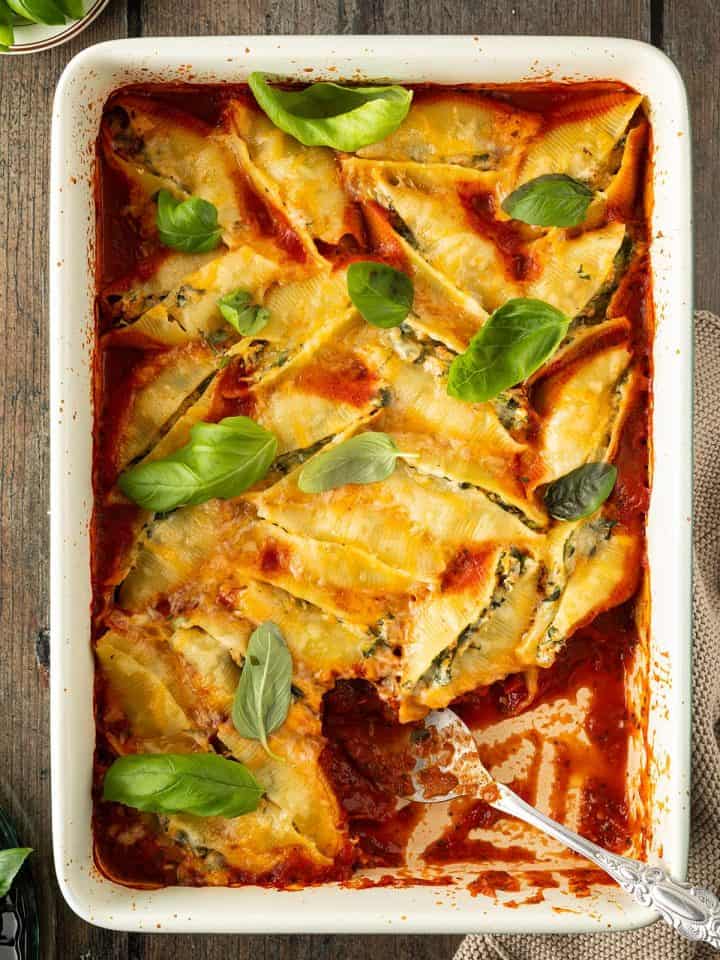 Eine Auflaufform mit gefüllten Muschelnudeln in Tomatensauce und mit frischem Basilikum, wo mit einem Löffel bereits eine Portion herausgenommen wurde.