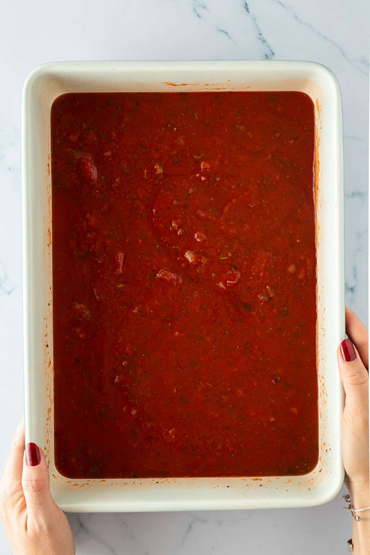 Eine Person hält eine große, rechteckige Auflaufform mit Tomatensauce in den Händen.