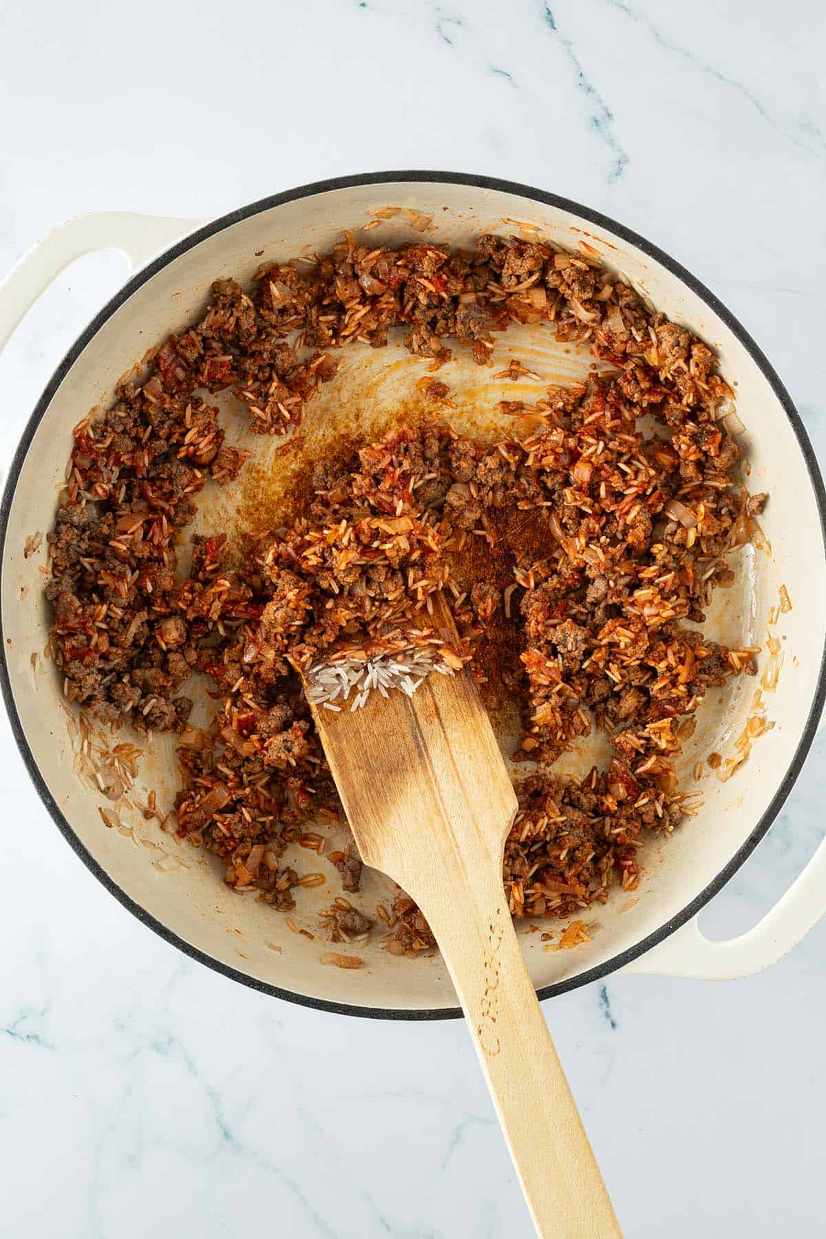 Angebratenes Hackfleisch mit Zwiebeln, Tomatenmark und ungekochten Reis in einer Pfanne.