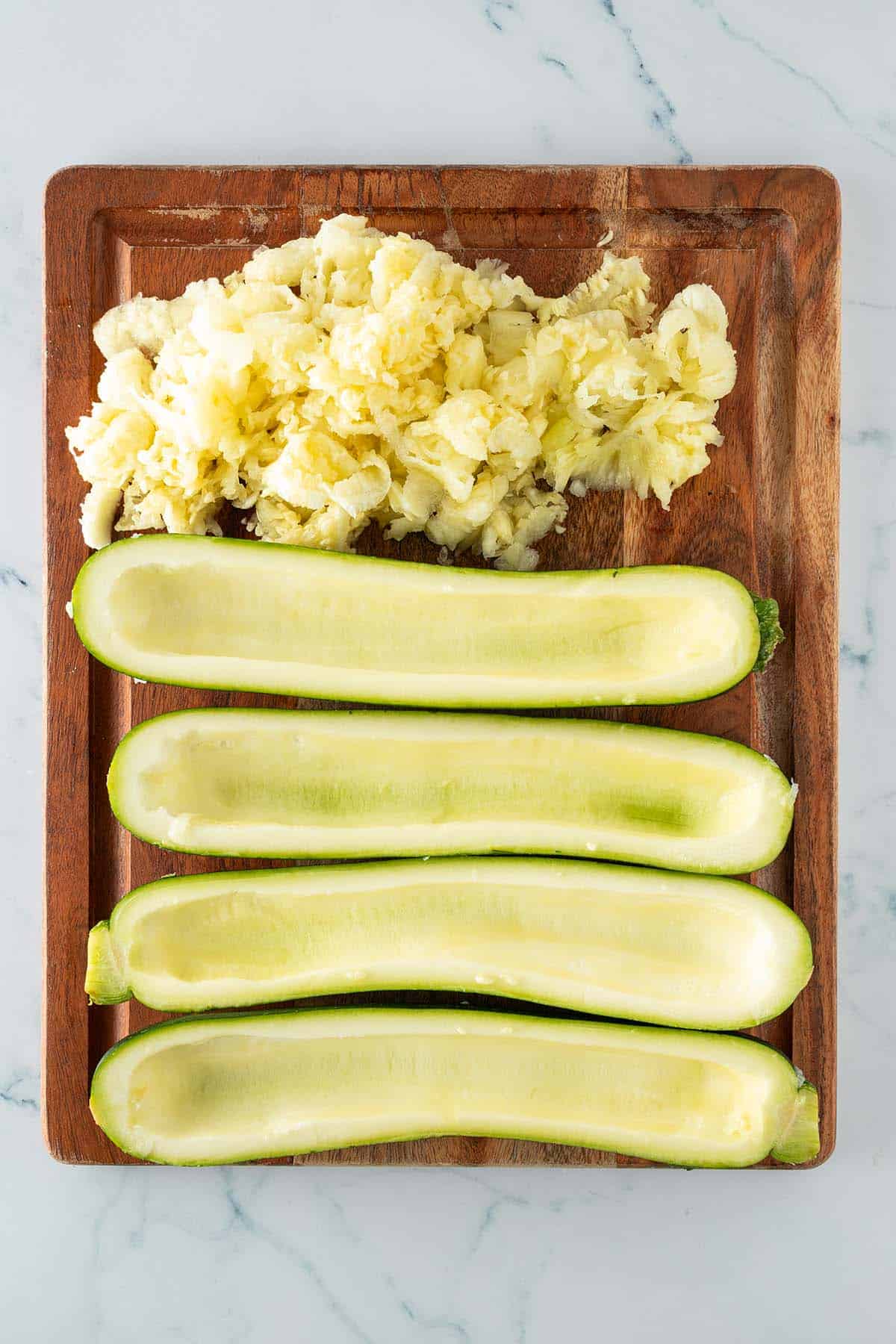 Ausgehöhlte Zucchinihälften und das entfernte Fruchtfleisch auf einem Holzbrett.