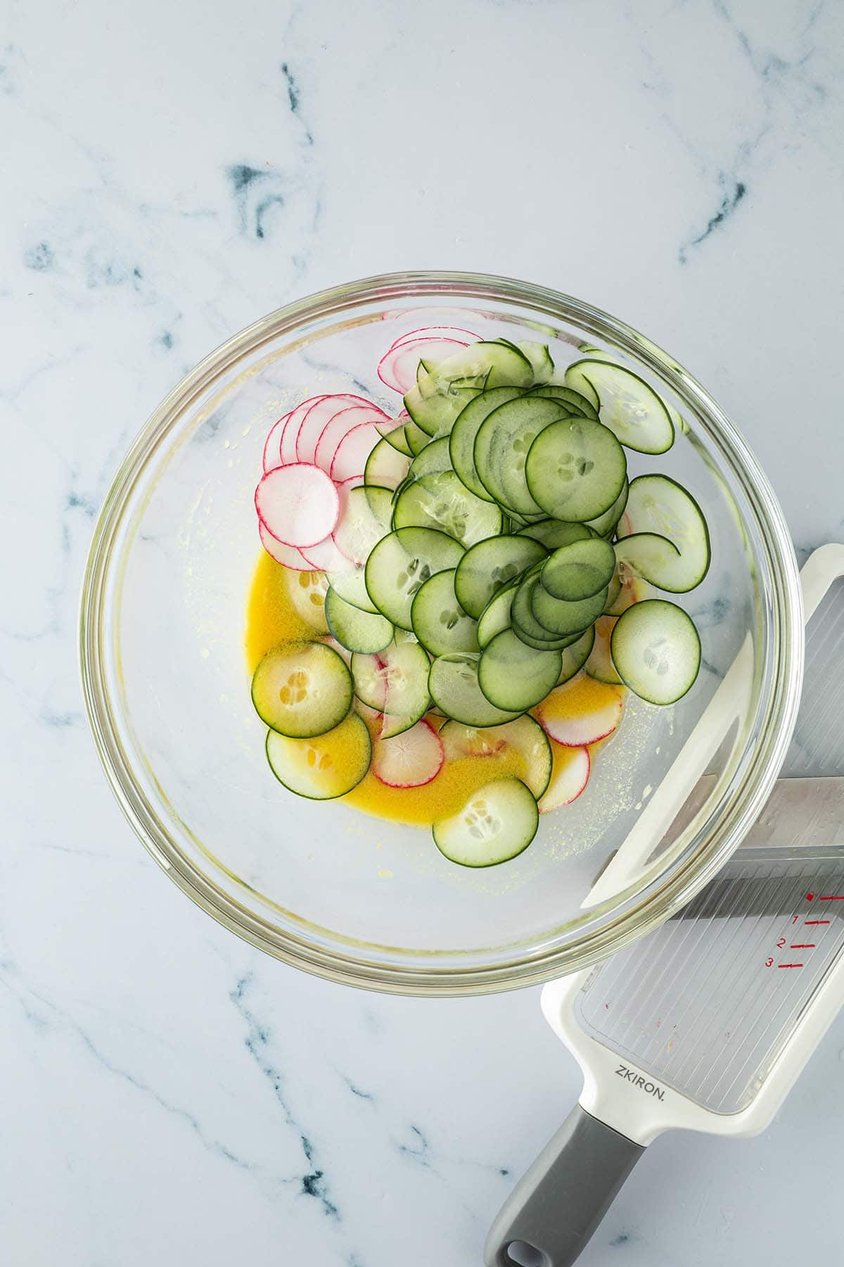 Eine Salatschüssel mit Dressing und in dünne Scheibchen gehobelte Radieschen und Gurken, daneben eine Gemüsehobel.
