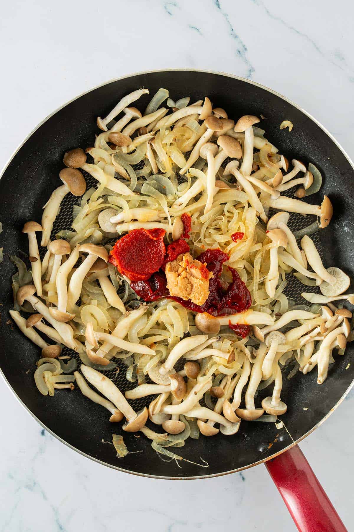 Pilze und Zwiebeln in einer Pfanne mit Gochujang, Miso und Tomatenmark vor dem Vermengen.