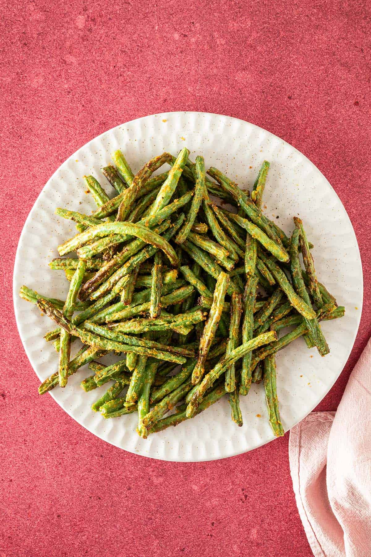 Grüne Bohnen, die in der Heißluftfritteuse zubereitetet wurden, auf einem weißen Teller, der auf einem roten Untergrund steht.