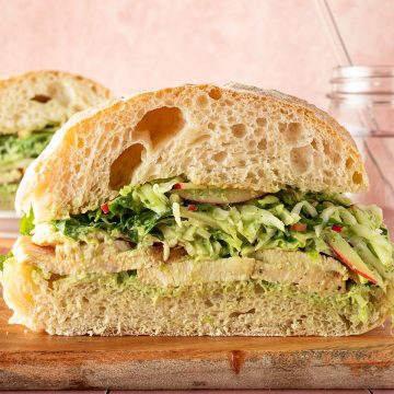 Aufgeschnittenes Ciabatta-Sandwich mit peruanischem H&auml;hnchen, Wei&szlig;kohl-Apfelsalat und cremiger gr&uuml;ner Sauce auf einem Holzbrett.