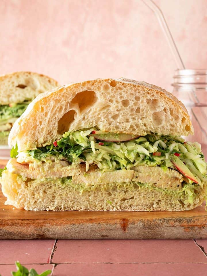 Aufgeschnittenes Ciabatta-Sandwich mit peruanischem Hähnchen, Weißkohl-Apfelsalat und cremiger grüner Sauce auf einem Holzbrett.