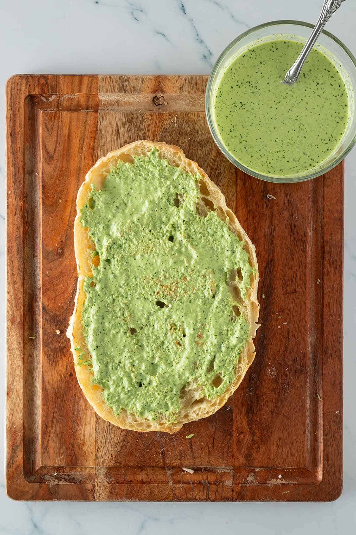 Ciabatta-H&auml;lfte mit Aji Verde bestrichen, daneben eine Schale mit der cremigen gr&uuml;nen Sauce auf einem Holzbrett.