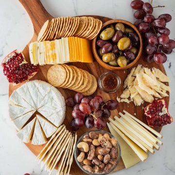 Käseplatte mit verschiedenen Käsesorten, Crackern, Trauben, Oliven, Nüssen und Granatapfel von oben fotografiert.