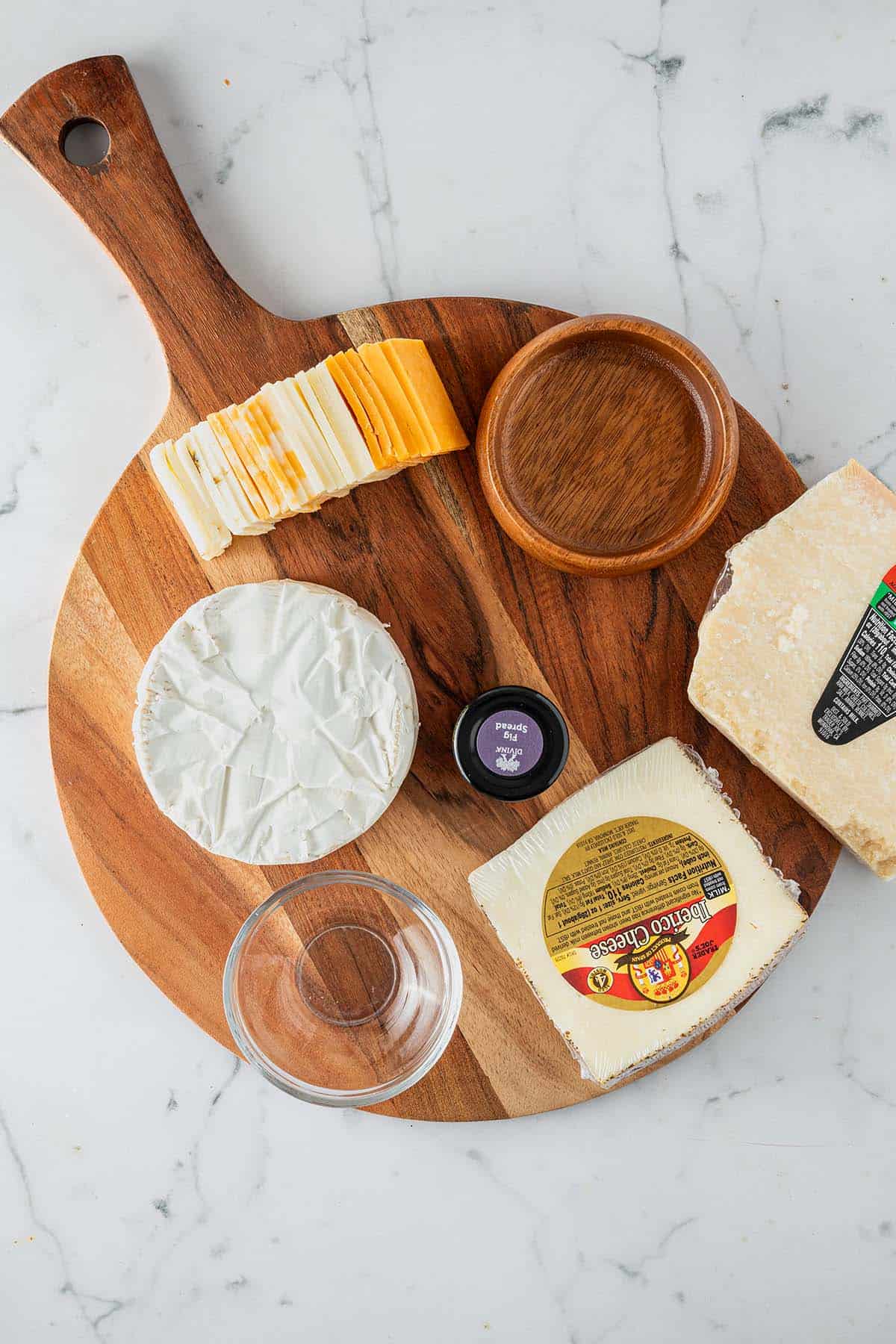 Holzbrett mit noch verpacktem Käse, Schälchen und vorbereitetem Zubehör für eine Käseplatte.
