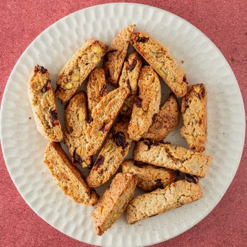 Teller mit goldbraun gebackenen Kardamom-Plätzchen mit Pistazien und Cranberries auf rotem Untergrund.