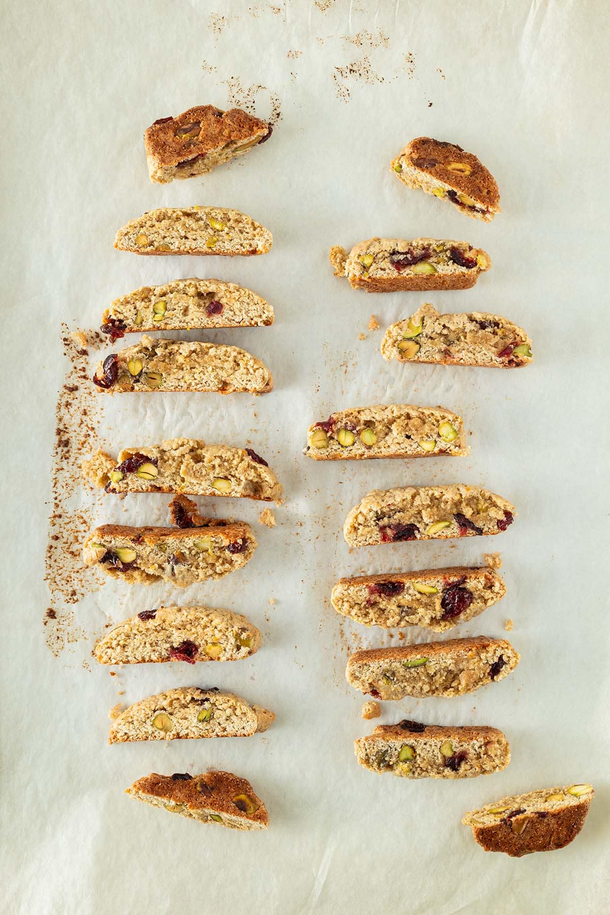 Aufgeschnittene Kardamom-Plätzchen mit Cranberries und Pistazien, gleichmäßig auf Backpapier verteilt.