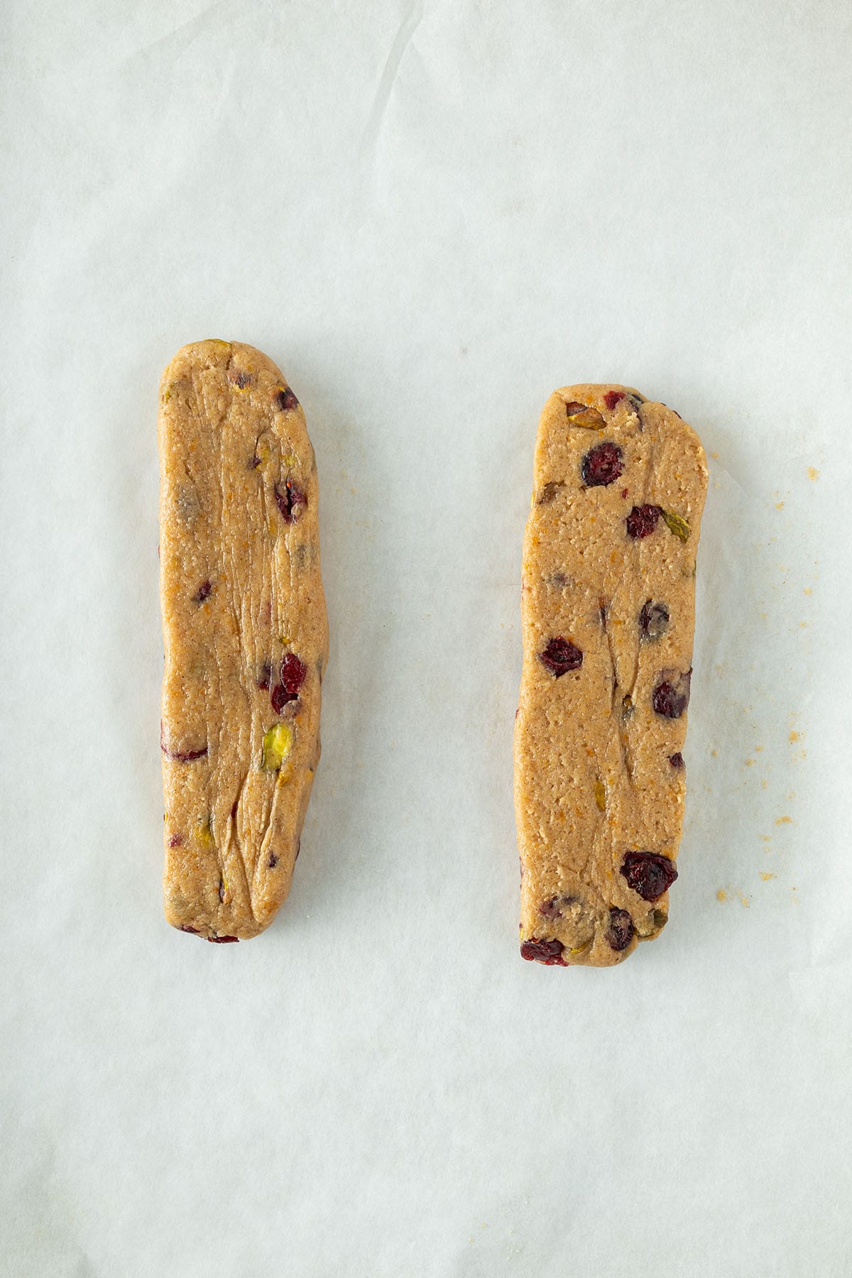 Zwei rohe Plätzchen-Teigstränge mit Pistazien und Cranberries auf Backpapier vorbereitet.
