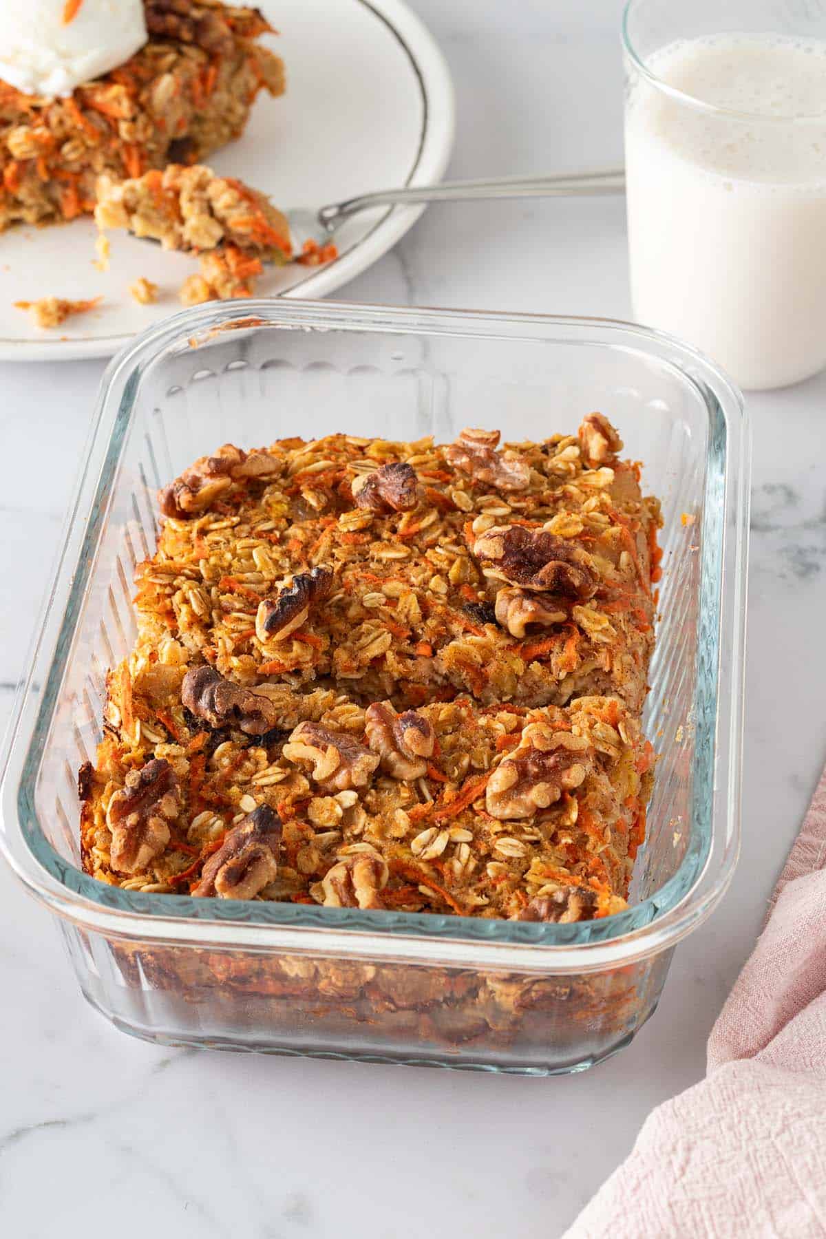 2 Portionen Karotten-Baked-Oats in einer Glasform, goldbraun gebacken mit Waln&uuml;ssen, eine Portion auf einem Teller mit einem Glas mit Milch im Hintergrund.