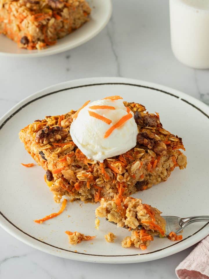 Ein Stück Karotten-Baked-Oats auf einem Teller, serviert mit Joghurt. Ein zweiter Teller mit einem Glas Milch im Hintergrund.